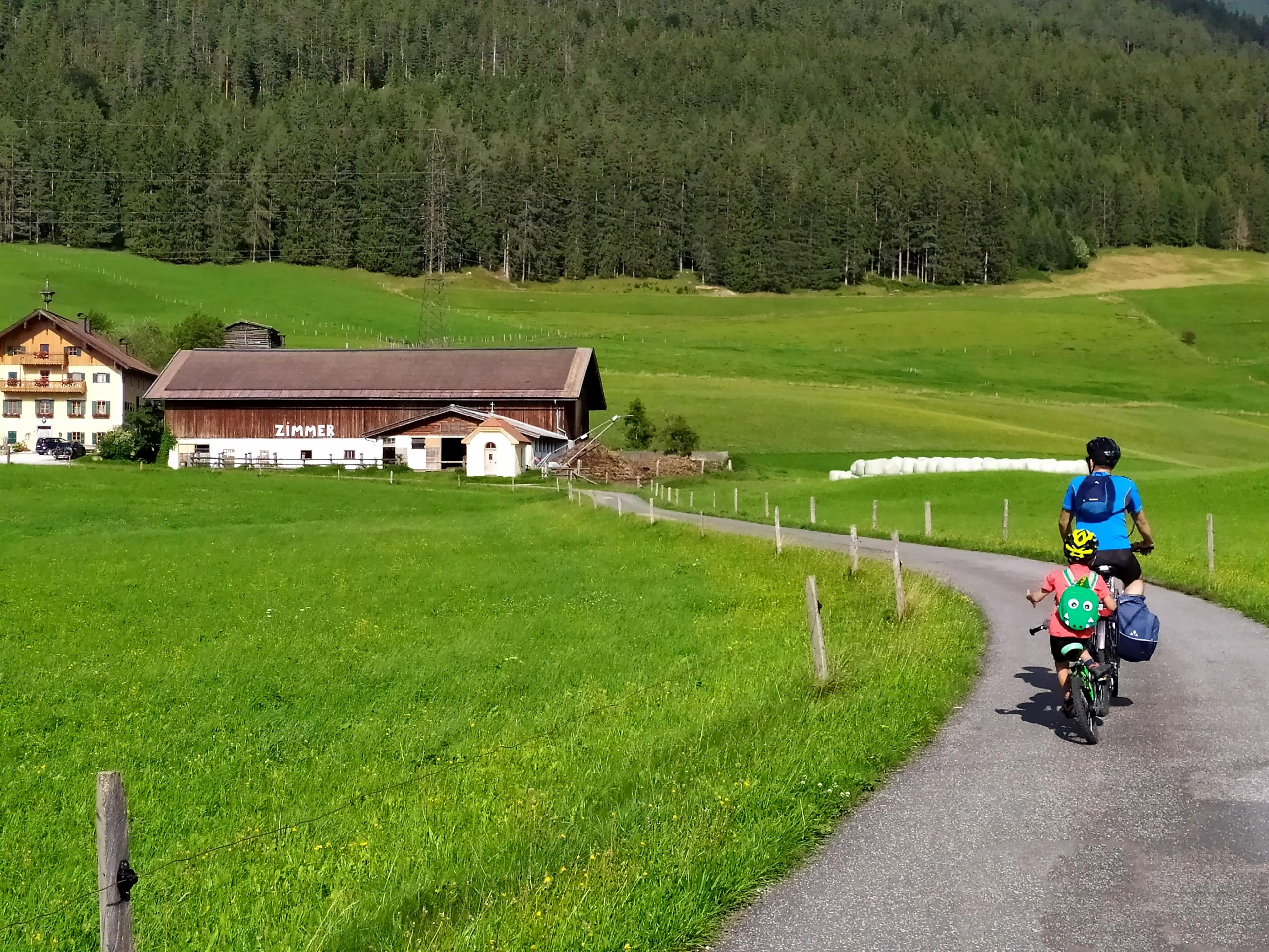 Famiglia in bicicletta lungo un percorso panoramico della Ciclabile dei Tauri, circondata da prati verdi e montagne.